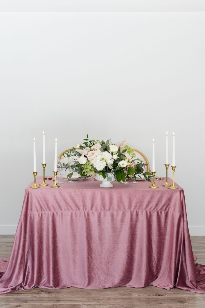 Mauve velvet table cloth adorned with gold candlesticks, gold taper candles, and a beautiful flower arrangement