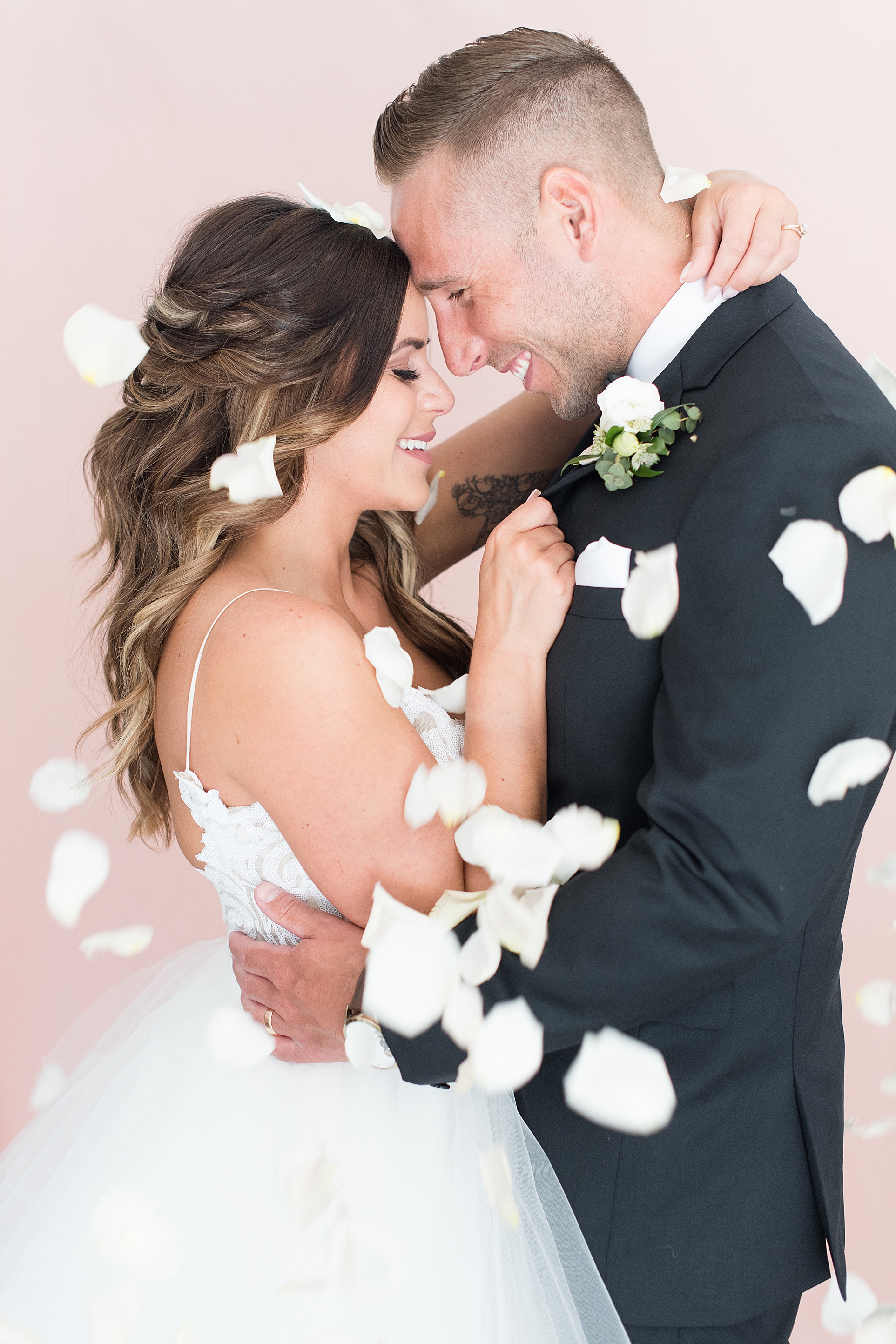 Bride and groom touch foreheads while white rose petals fall around them