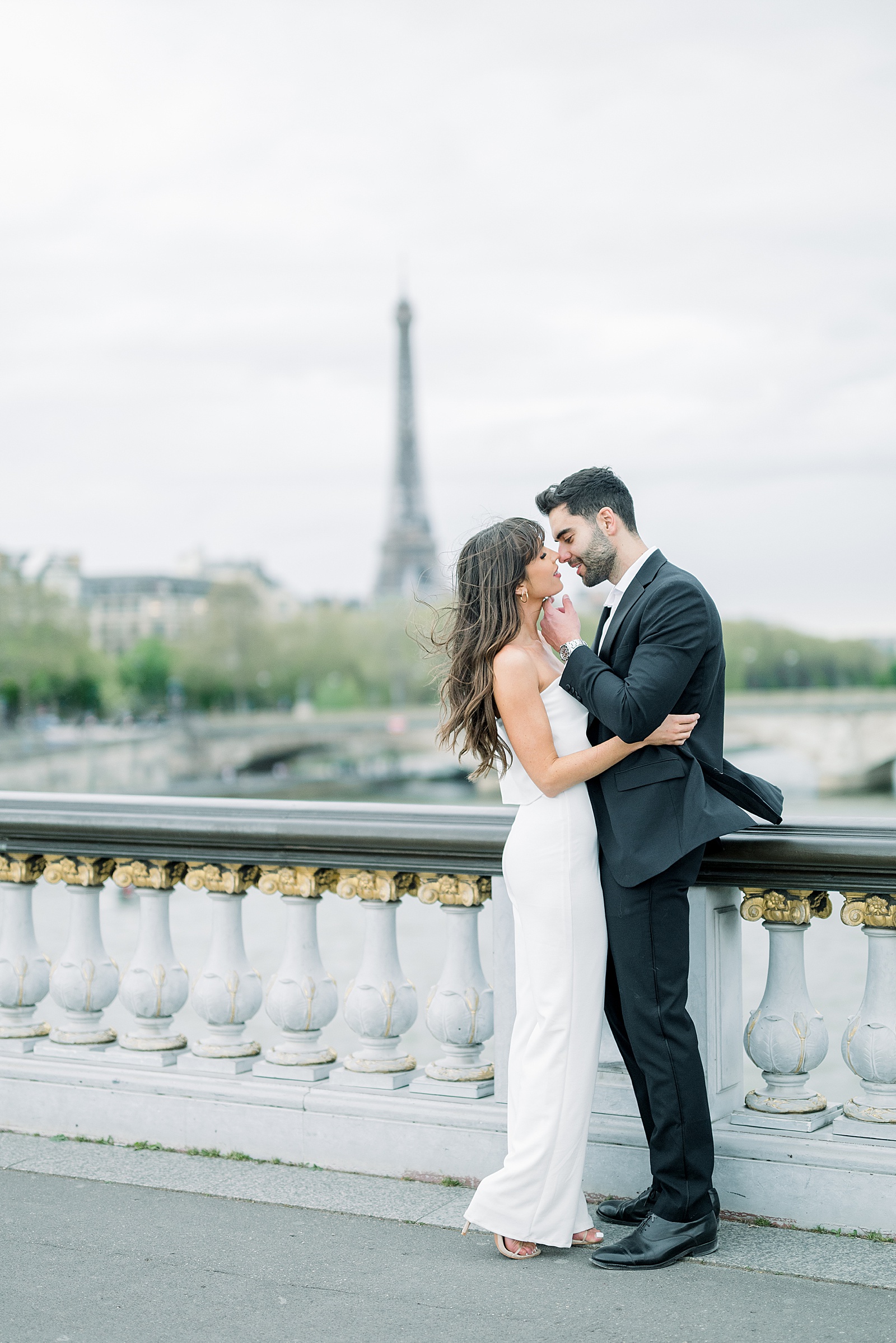 A couple stands on the pont alexandre iii on a windy day with the Eiffel tower in the background
